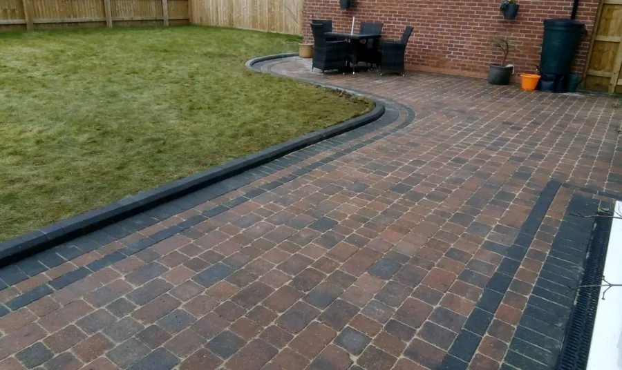 New block paving with a dark edge course curving round a lawn by a brick garage